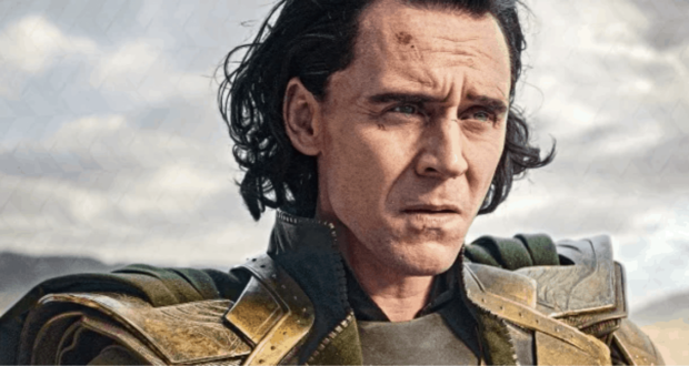 A person with long dark hair and pale skin wears a green and gold armored costume, looking serious against a cloudy sky background.