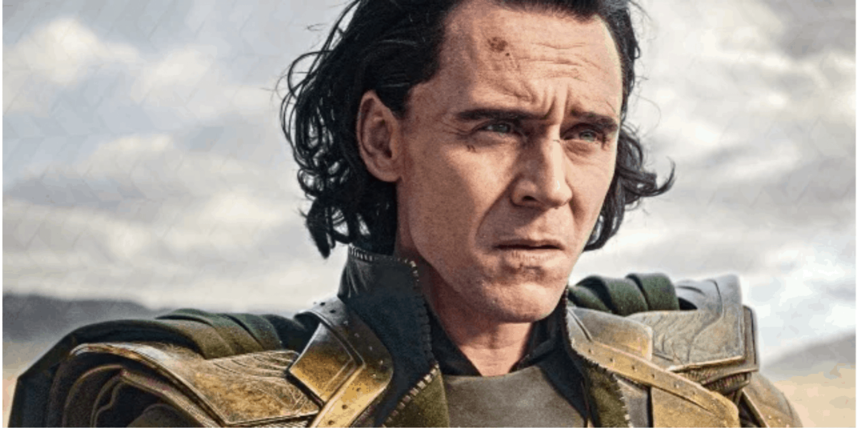 A person with long dark hair and pale skin wears a green and gold armored costume, looking serious against a cloudy sky background.