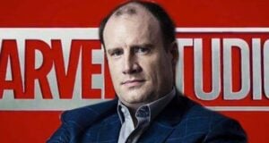 A man in a suit poses in front of a large red and white Marvel Studios logo background.