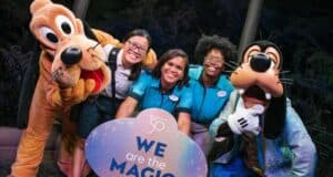 A group photo of three Disney cast members standing with Pluto and Goofy. The smiling cast members are holding a circular sign that reads, "We are the magic." They are outdoors, with trees and lights in the crisp background, truly showing what it means to be part of the Disney enchantment.