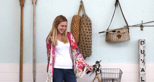 A woman with long blonde hair, wearing a white shirt and floral kimono, smiles while standing next to a vintage bicycle against a light blue wall adorned with nautical items inspired by Disney Resort Decor.