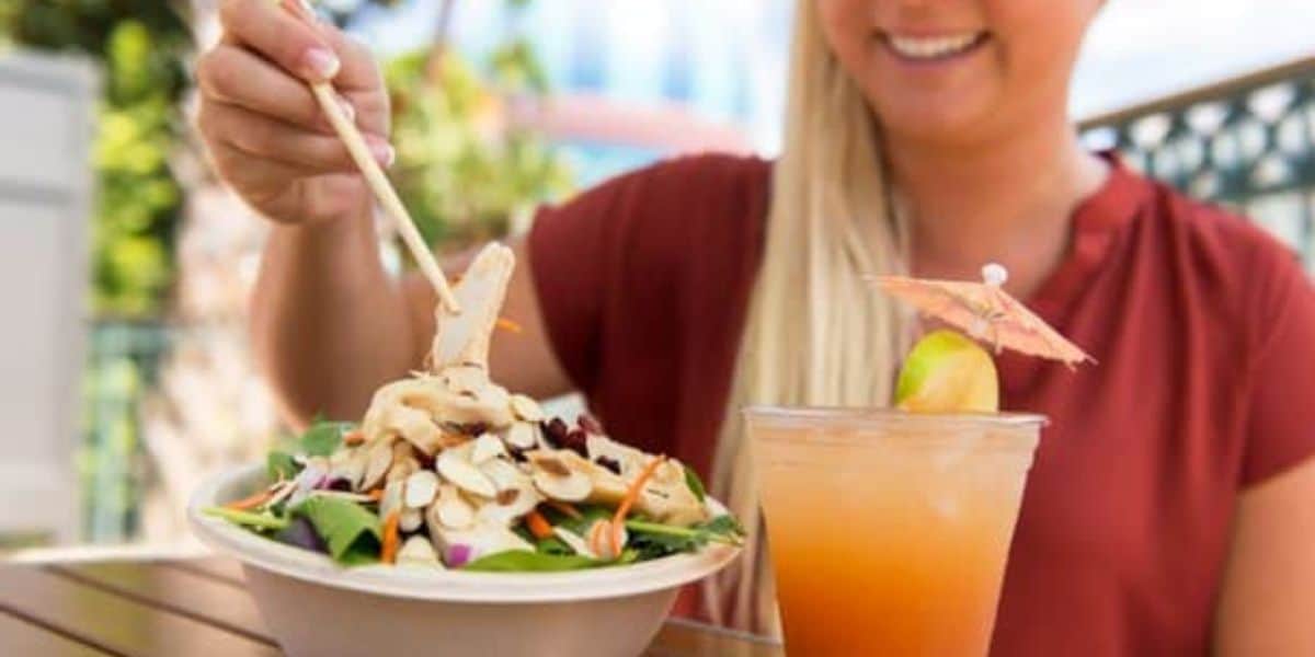A person in a red shirt is enjoying healthy eating with chopsticks, about to savor a salad topped with sliced almonds in a white bowl. Next to the bowl is a tropical drink with a small umbrella, bringing a touch of Walt Disney World magic to their dining experience.
