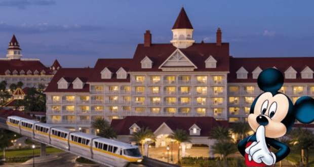 At dusk, a large deluxe hotel with a red roof and white exterior glows with warm lighting. A monorail glides past, embodying the charm of a Disney Resort. In the foreground, an animated character stands thoughtfully with a finger to their mouth.