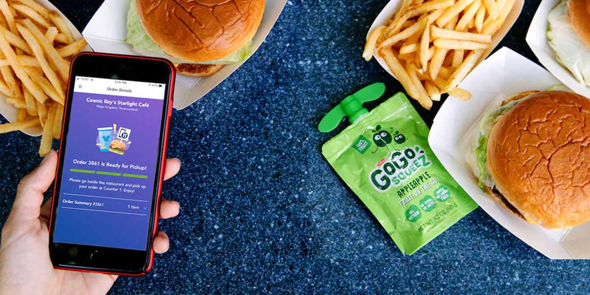 A hand holds a phone displaying a Disney World Mobile Ordering app next to trays of burgers, fries, and a pouch of GoGo squeeZ apple sauce on a blue tabletop.