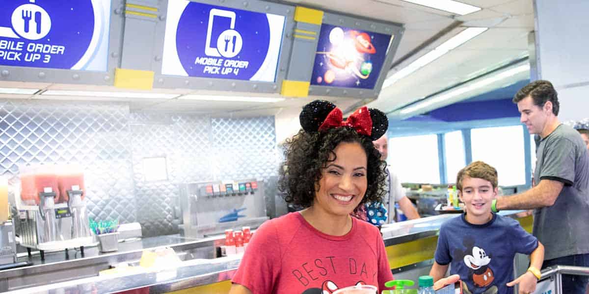 A smiling woman wearing Mickey Mouse ears and a “Best Day Ever” shirt carries food trays in a modern restaurant, with a boy beside her and Disney World Mobile Ordering pick-up signs glowing in the background.