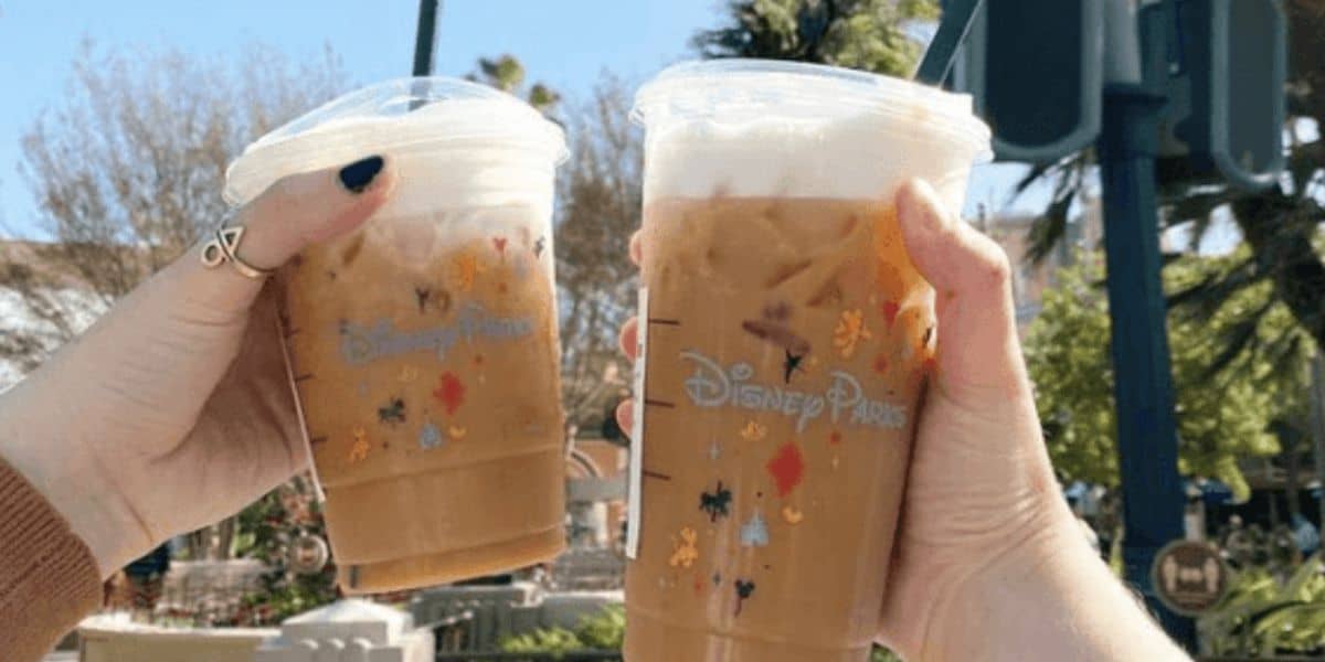 Two hands holding iced coffee cups with Disney Parks logos and playful Mickey Mouse silhouettes reflect the excitement of a sunny day. Could these refreshing delights be part of the elusive Disney Starbucks secret menu? Trees and a blurred green sign complete this magical moment.