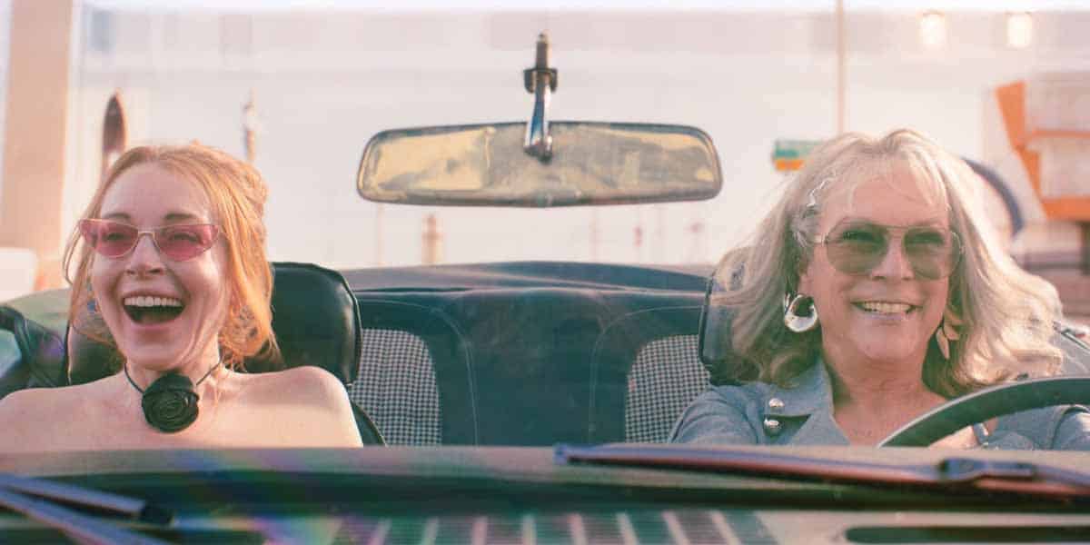 Two women wearing sunglasses laugh and smile while riding in a convertible car on a sunny day, sharing Mother's Day joy. The driver has gray hair, and both seem to be enjoying themselves as if cruising to Disney World.