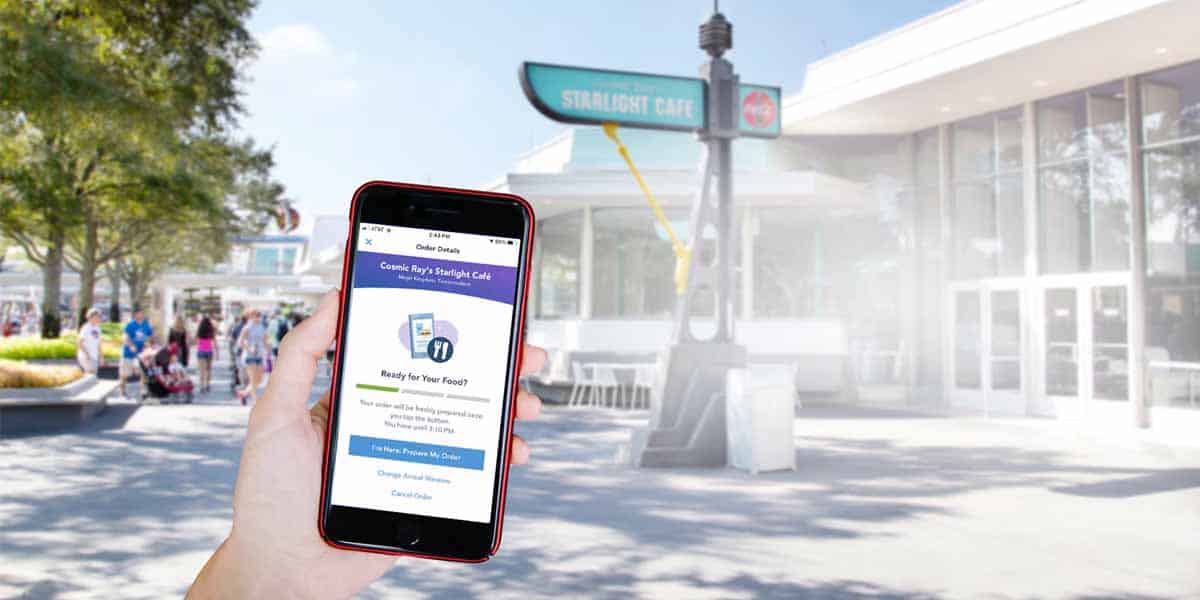 A person holds a smartphone displaying a Disney World Mobile Ordering screen in front of Starlight Cafe, with people and trees visible in the background.