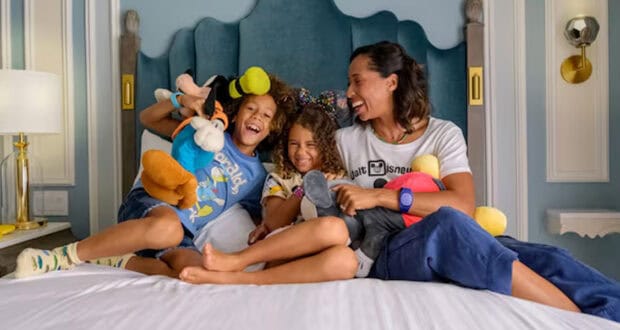 A woman and two children sit on a bed, smiling and laughing while holding stuffed cartoon toys, enjoying a cozy Disney Staycation in a well-decorated bedroom.