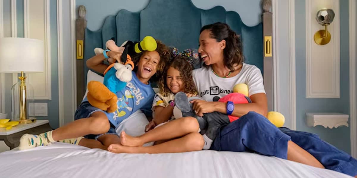 A woman and two children sit on a bed, smiling and laughing while holding stuffed cartoon toys, enjoying a cozy Disney Staycation in a well-decorated bedroom.