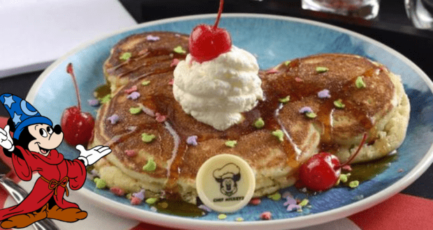 A plate features three pancakes arranged in the shape of a famous mouse's head, topped with whipped cream and a cherry. Colorful sprinkles decorate the pancakes, alongside cherries and a white chocolate piece with a chef hat design. It's the perfect Mickey snack for any Disney Dining experience, complete with a wizard character on the left.