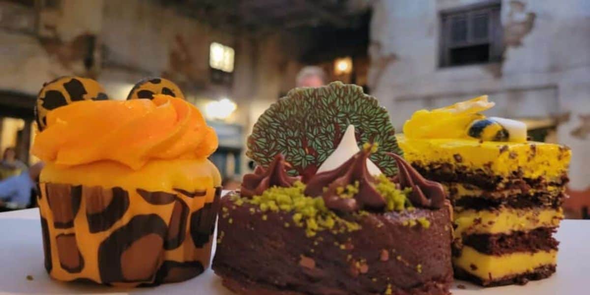 A close-up of three Disney World desserts. On the left, a giraffe-patterned cupcake with orange frosting and chocolate spots. In the center, a Tusker House-inspired chocolate cake with a decorative tree. On the right, a layered yellow and chocolate cake with lemon garnish.