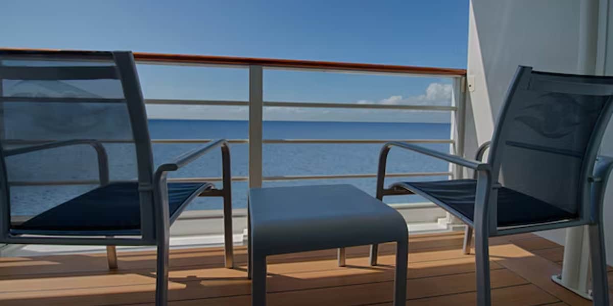 Two empty chairs and a small table sit on the verandah of a Disney Cruise Verandah Stateroom, overlooking a calm blue ocean, with a clear sky and a few clouds in the distance.