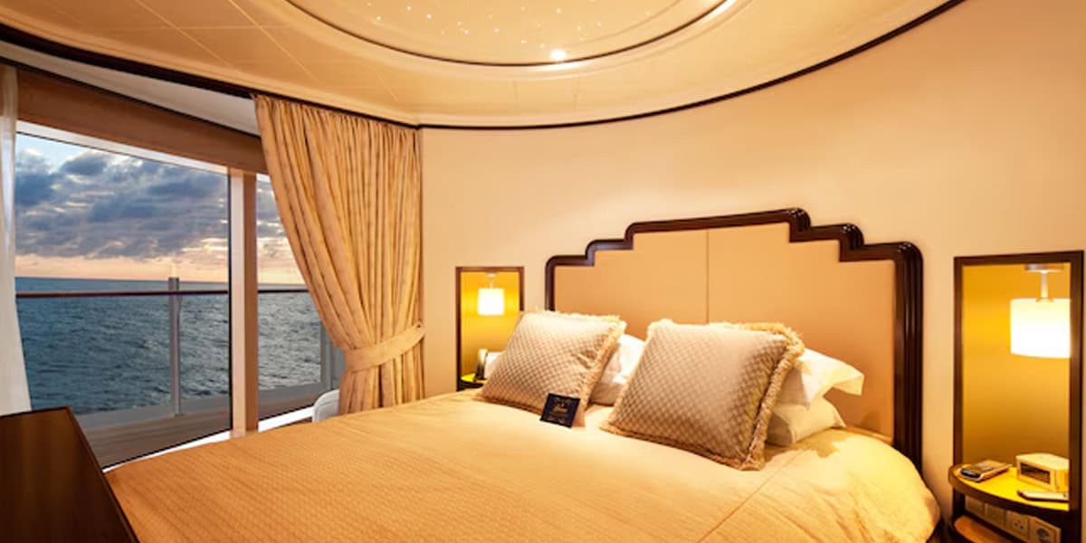 A luxurious Disney Cruise Verandah Stateroom bedroom with a large bed, elegant headboard, plush pillows, and warm lighting. Floor-to-ceiling windows with beige curtains reveal an ocean view at sunset.