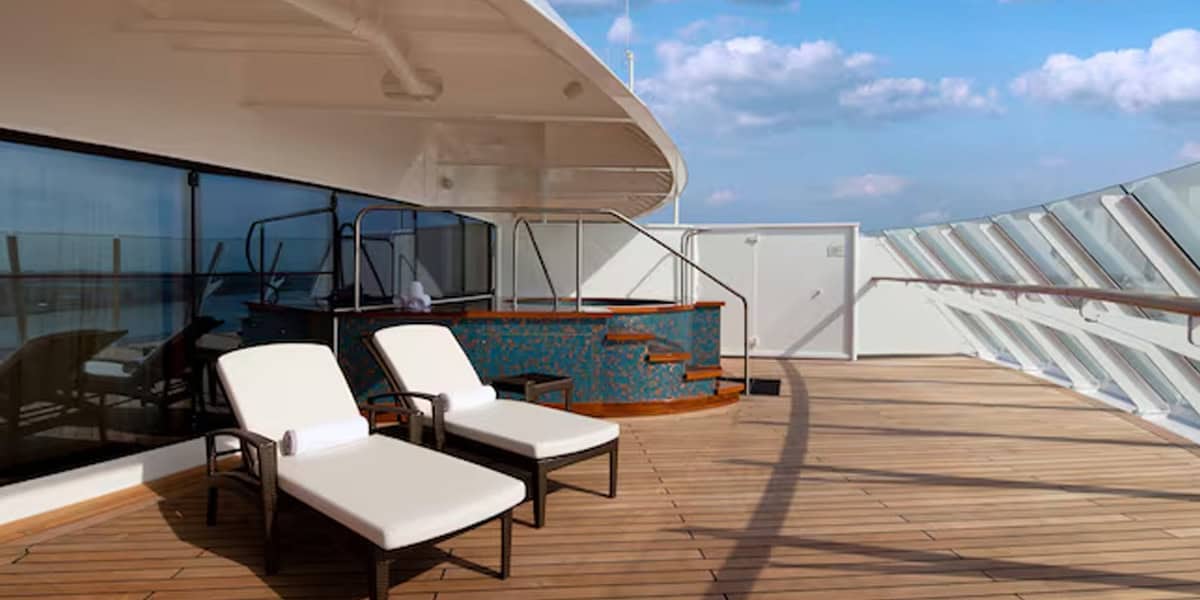 Two cushioned lounge chairs sit on the spacious wooden deck of a Disney Cruise Verandah Stateroom, next to a small mosaic-tiled hot tub. Glass panels line the deck, with a blue sky and clouds visible above.