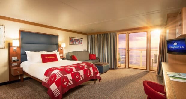 A bright, spacious hotel room styled like a Disney Cruise Verandah Stateroom, featuring a large bed with a red nautical-themed blanket, gray sofa, desk with TV, and glass doors opening to a balcony with an ocean view at sunset.