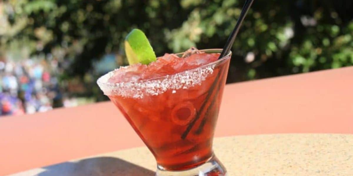 A red cocktail garnished with a lime wedge sits in a salt-rimmed glass on a sunny outdoor table, hinting at affordable drink prices. A black straw rests in the drink, with a blurred natural backdrop of greenery in the distance.