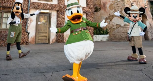 Goofy, Donald Duck, and Mickey Mouse are dressed in safari outfits, enthusiastically posing outdoors in front of a textured stone building at Tusker House Restaurant. Donald stands in the center with arms wide open, while Goofy and Mickey strike welcoming poses.