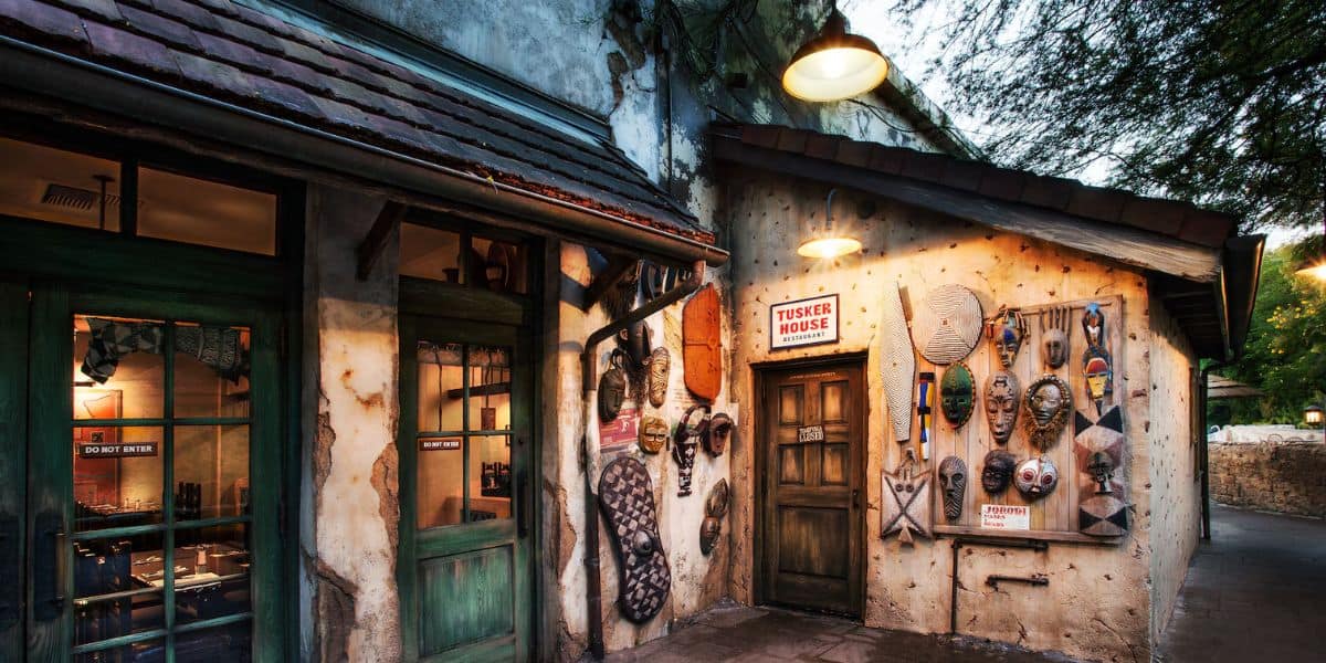 The rustic exterior of the building, adorned with African masks and a sign reading "Tusker House Restaurant" above the wooden door, exudes charm. Warm light from an overhead lamp complements the scene, with trees gently swaying in the background.