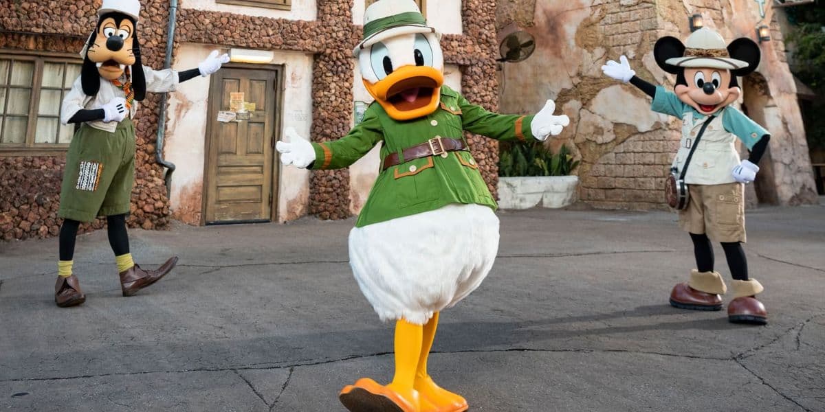 Goofy, Donald Duck, and Mickey Mouse are dressed in safari outfits, enthusiastically posing outdoors in front of a textured stone building at Tusker House Restaurant. Donald stands in the center with arms wide open, while Goofy and Mickey strike welcoming poses.