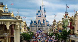 A large crowd gathers on Main Street in front of a blue-and-white fairytale castle at a theme park, surrounded by colorful buildings and American flags—unaware of the hidden utilidors bustling beneath them—under a partly cloudy sky.