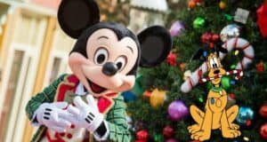 Mickey Mouse in festive holiday attire stands by a decorated Christmas tree, with Pluto holding a candy cane in his mouth, surrounded by Christmas lights—perfect for adding Disney holiday magic to your home or as a charming free gift.