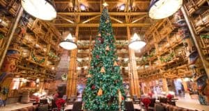 A grand lodge decorated for the holidays, featuring a towering tree adorned with lights, ornaments, and festive ribbons. The wooden interior, reminiscent of Walt Disney World's magic, is lit by chandeliers and surrounded by lush garlands and intricately carved pillars.