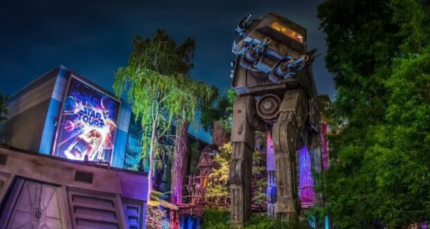 A nighttime view of a Star Wars-themed attraction at Walt Disney World features a large, illuminated AT-AT walker among trees. A brightly lit "Star Tours" sign is visible on a building to the left. The scene is enhanced with colorful lighting effects, showcasing the magic of Disney attractions.
