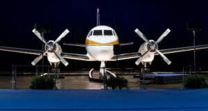 A twin-propeller airplane with a yellow stripe is displayed indoors on a stage at one of the Disney museums, surrounded by blue carpeting and green plants, under spotlights against a dark curtain backdrop.