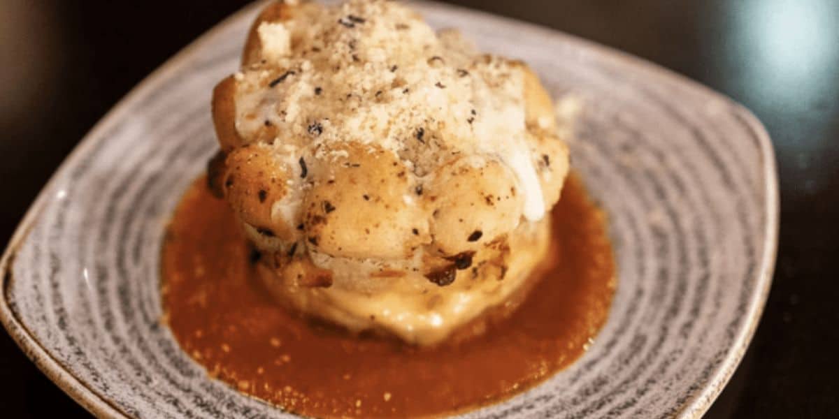 A gourmet dish featuring a layered stack of cooked ingredients topped with sauce and grated cheese, served on a textured plate with a brown sauce spread on the base. This elegant presentation is paired with crispy garlic bread, creating a delightful contrast against the dark background.