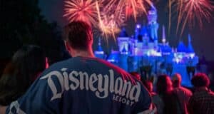 A group of people watches a colorful fireworks display at night. In the foreground, a person wearing a "Disneyland Resort" shirt stands with their back to the camera during Sweethearts Nite at Disney. In the background, a Disney castle is beautifully illuminated.