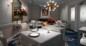 A cozy, elegant dining room features tables with white tablecloths set for two, a beautifully decorated fireplace, ornate chandelier, and floral arrangements. A portrait hangs above the fireplace, and soft lighting creates a warm ambiance—the perfect spot to enjoy My Favorite Eats Money Can Buy at Walt Disney World.