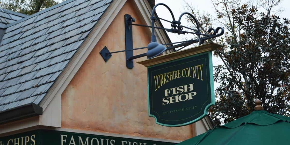 A small building with a pitched roof features a green and gold sign that reads "Yorkshire County Fish Shop." Below, another portion of the sign advertises "Famous Fish and Chips," one of My Favorite Eats Money Can Buy at Walt Disney World. Trees and an umbrella are visible in the background.