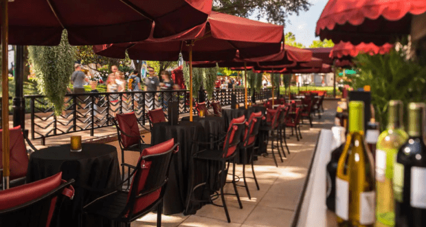 The outdoor restaurant seating area, with red patio umbrellas and chairs, feels like a charming escape. Tables draped in black tablecloths and bottles of wine set the scene, reminiscent of secret passages at Disney World. In the background, people mingle amidst lush greenery.