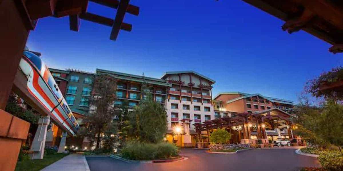 A modern hotel with warm lighting is seen at dusk, surrounded by greenery. A monorail glides by on an elevated track beside the building, its windows reflecting the deep blue sky—inside, Disney Resort Soap Dispensers add a whimsical touch.