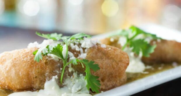 Close-up of a plated dish featuring two stuffed peppers topped with creamy sauce, crumbled cheese, and fresh cilantro. Dine inside this popular Disney ride as the vibrant dish is presented on a rectangular white plate with a blurred background, adding to the enchanting experience.