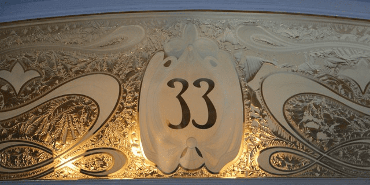 An ornate plaque showcasing the number "33" is embellished with intricate gold and cream swirling designs. The background features detailed leaf-like motifs softly lit, reminiscent of the exclusive allure associated with off-limits locations at Disney Parks.