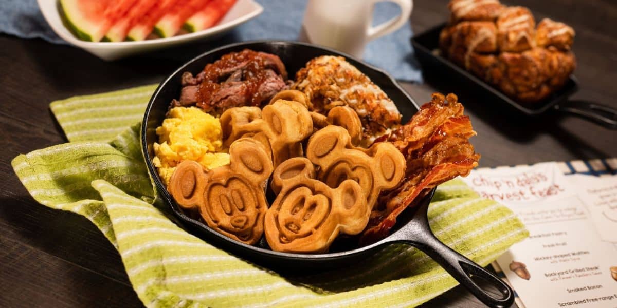 A skillet filled with Mickey Mouse-shaped waffles, scrambled eggs, bacon, and sausage sits on a green striped cloth at Garden Grill Restaurant. In the background, there's watermelon, a pastry, and a small pitcher of milk, with a Disney menu partially visible on the side.