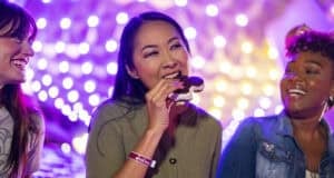 Three women smile and laugh under string lights, with one woman in the center holding a large s’more—sharing tips from their Disney vacation packing list in a warmly lit, festive atmosphere.