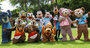 A group of Disney characters, venturing outside of parks, don vibrant costumes as a blue creature and animated animals like a mouse, duck, dog, and bears pose cheerfully on a grassy area with palm trees swaying in the background.