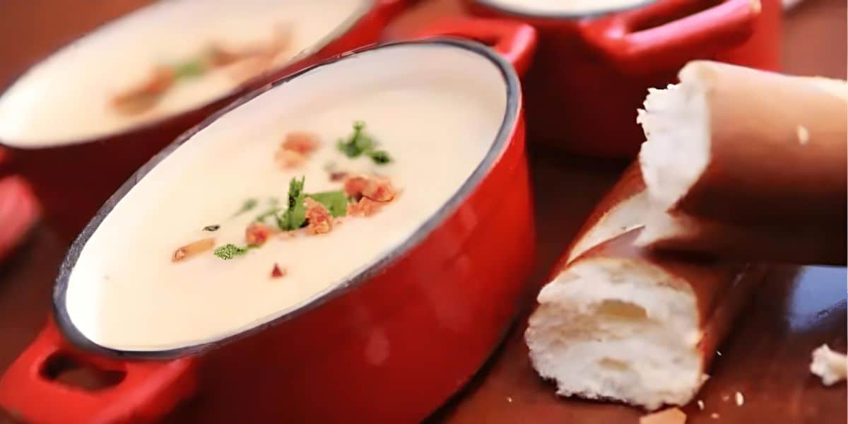 A close-up of creamy soup, resembling the weird soups at Disney World, in red bowls, garnished with herbs and crispy bits. Next to the bowls is a partially broken baguette resting on a wooden surface, highlighting the texture and detail of both soup and bread.
