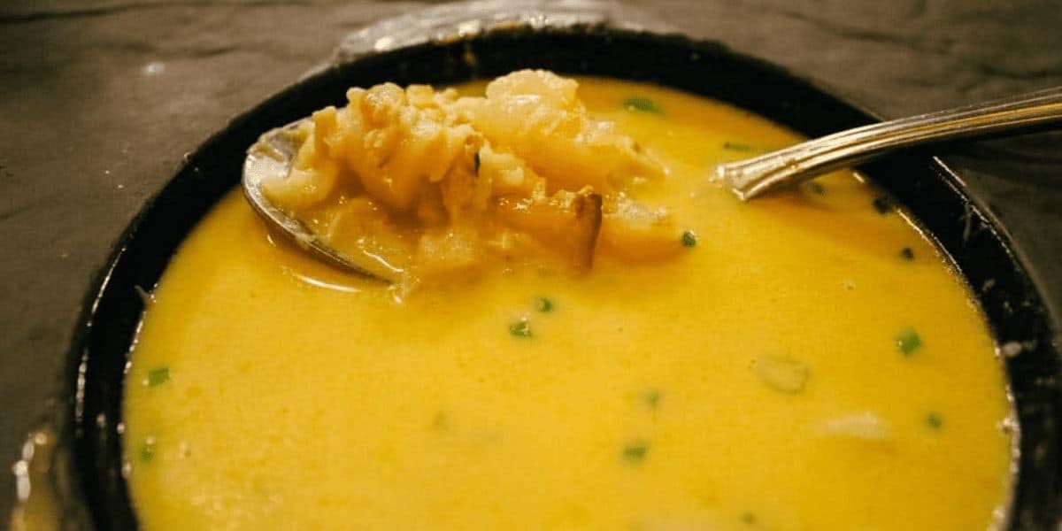 A bowl of creamy yellow soup containing pieces of cauliflower and garnished with green herbs, reminiscent of the weird soups at Disney World. A spoon rests inside the bowl, lifting some of the soup and ingredients.