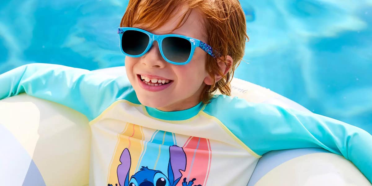 Smiling child with red hair wearing blue sunglasses and a colorful Stitch rash guard lounges in a pool on a white inflatable float—perfect for your Disney vacation packing list.