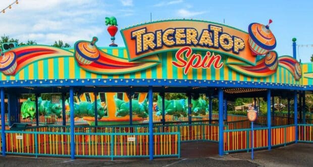 A colorful amusement park ride entrance, "TriceraTop Spin," features dinosaur-themed vehicles and vibrant green, orange, and yellow decorations. While enjoying the blue sky and trees in the background, consider this one of the rides to skip at Animal Kingdom if you're short on time.