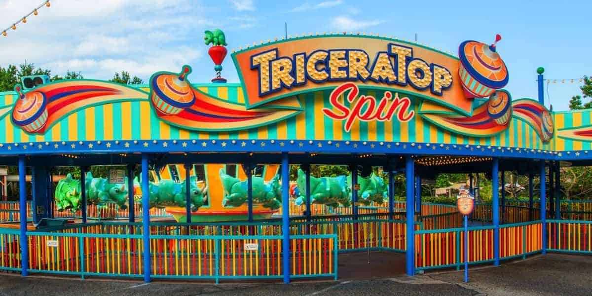 A colorful amusement park ride entrance, "TriceraTop Spin," features dinosaur-themed vehicles and vibrant green, orange, and yellow decorations. While enjoying the blue sky and trees in the background, consider this one of the rides to skip at Animal Kingdom if you're short on time.