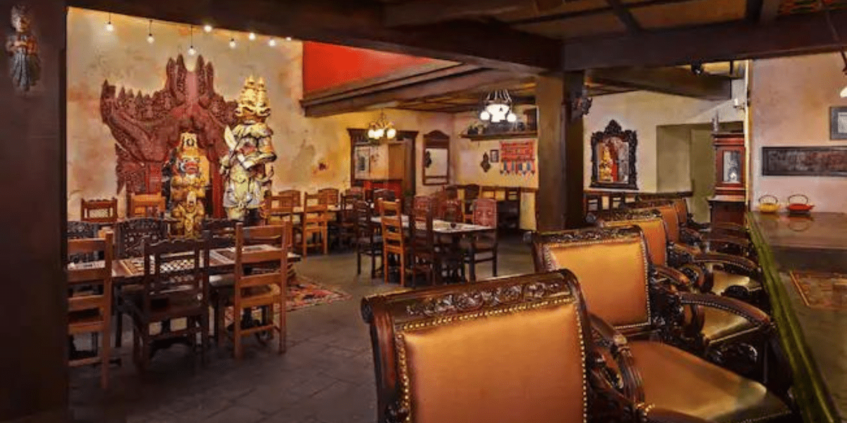 Nestled like an undiscovered gem among underrated restaurants at Disney World, this cozy interior features wooden tables, ornate carved leather seats, and rustic decor. Dim lighting and vibrant wall art, including colorful masks and paintings, craft a warm and inviting atmosphere.