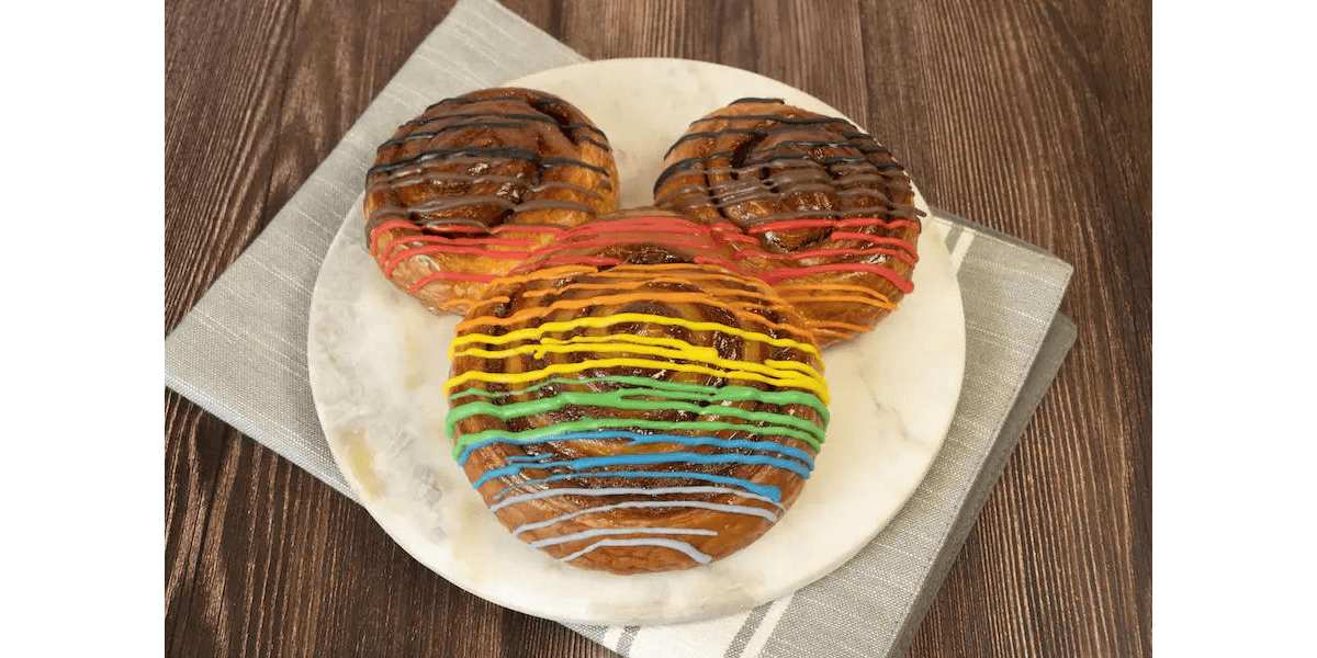 A pastry shaped like a popular mouse character is on a marble plate atop a gray napkin, celebrating Pride with three round, connected sections adorned in vibrant icing stripes of red, orange, yellow, green, blue, and purple.