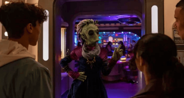 A group of people stand at the entrance of a futuristic room reminiscent of Star Wars: Galactic Starcruiser. An extraterrestrial figure dressed in colorful attire gesticulates while facing them, set against a backdrop of vibrant lights and sleek, sci-fi interiors.