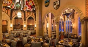 An elegant, medieval-themed dining hall in the heart of Magic Kingdom park features arched stone doorways, numerous chandeliers, and colorful flags hanging from the ceiling. Tables are set with yellow napkins and cushioned seating, creating a warm, inviting atmosphere for the best dining experience.