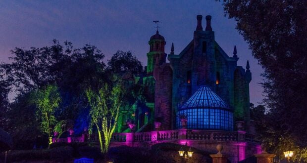 A spooky, dark mansion is lit with eerie green, purple, and blue lights against a twilight sky. Surrounded by trees, the building evokes Disney's Haunted Mansion ride with its distinct glass conservatory, towers, and chimney stacks—perfect for a haunted mansion trivia night.